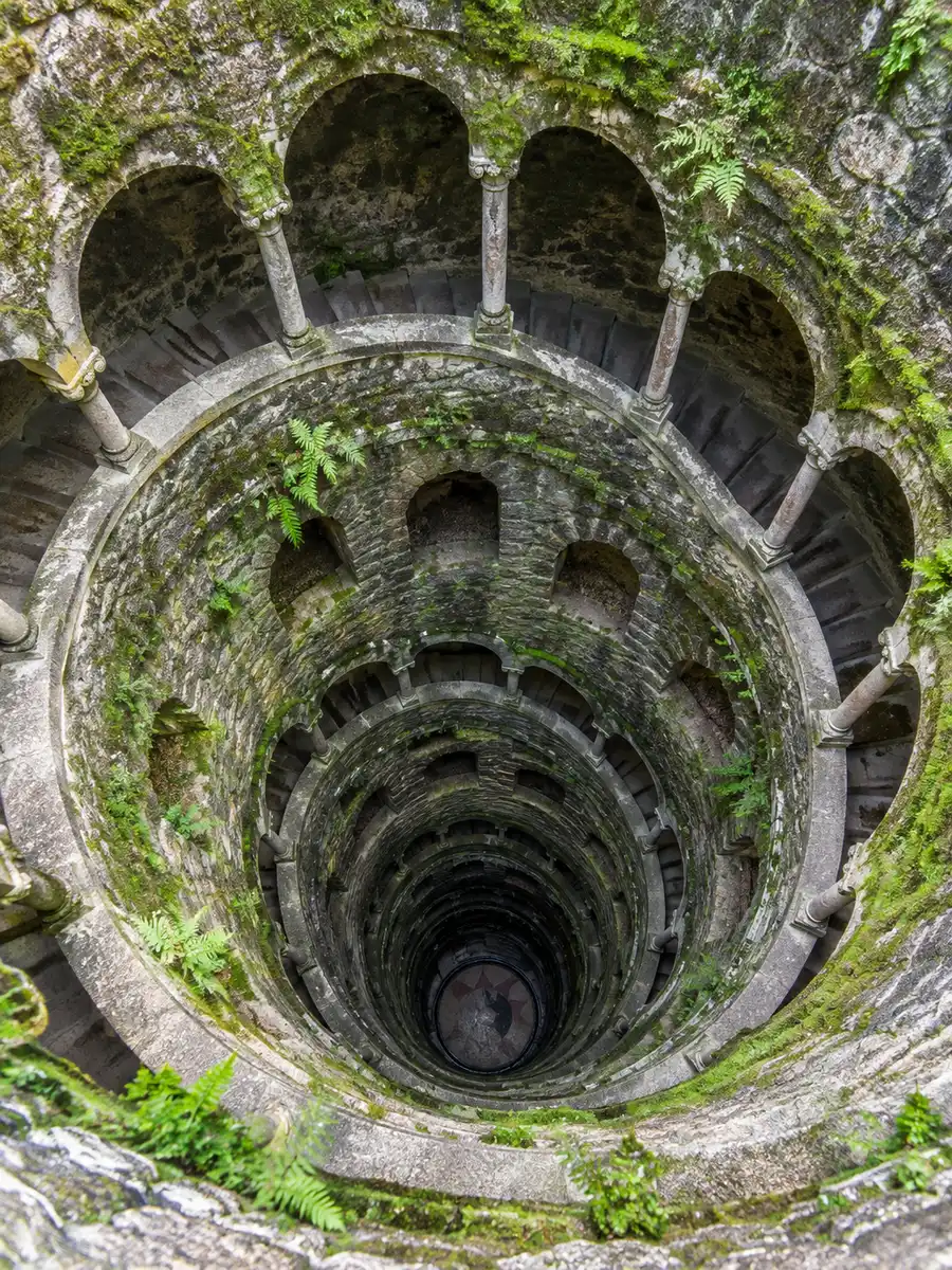 Looking down the spiral staircase of the Initiation Well at Quinta da Regaleira, with moss on the stone arches